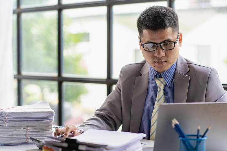 Person sitting at a desk looking at a laptop with a stack of reports next to them